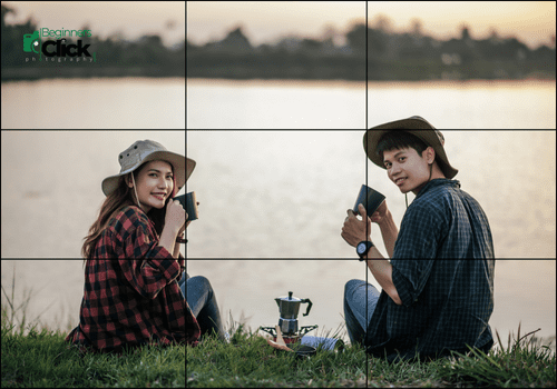 A young man and woman sitting by a lakeside during sunset, holding mugs and smiling at the camera. A coffee brewing set is placed on the grass in front of them.