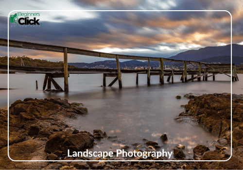 Serene Horizons – Landscape Photography A long-exposure shot of a calm water body with a wooden pier stretching into the distance, mountains in the background, and labeled “Landscape Photography” with the Beginners Click Photography logo.