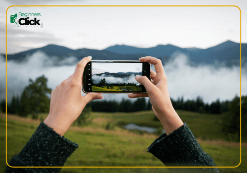 Capturing Nature Through a Phone Lens A person holding a smartphone to photograph a misty mountain landscape, with green hills and trees in the foreground, and the "Beginners Click Photography" logo in the top-left corner.