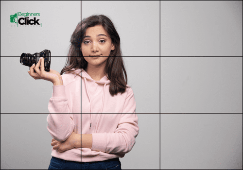 Beginner Photographer Learning Composition Rules A young woman in a pink hoodie holding a camera in her right hand, standing against a plain gray background. The image has a rule-of-thirds grid overlay.