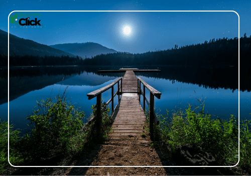 Moonlit Tranquility at the Lake A wooden dock extends into a calm lake under the moonlight, surrounded by mountains and forest, with clear reflections on the water.