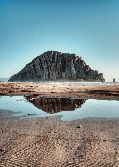 Reflections of Progress Photo of a large rock formation reflected in a tide pool on a beach, taken during a beginner's journey of learning photography from scratch, capturing symmetry, texture, and natural light.